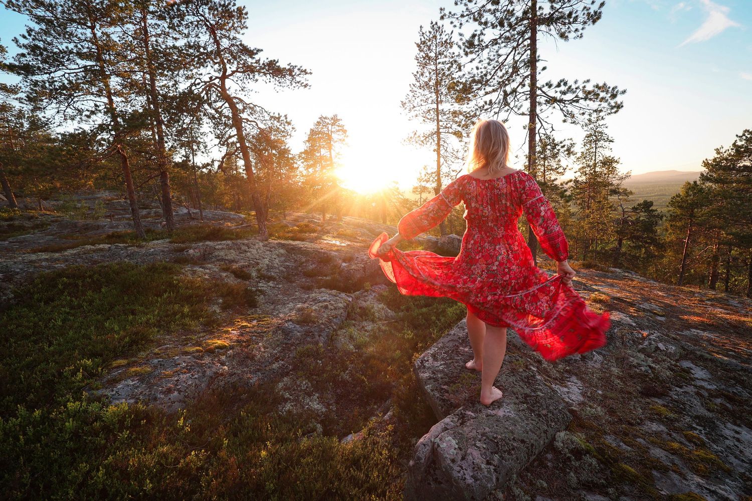 Woman in red floral dress standing on rocky outcrop watching golden sunset through pine trees.
