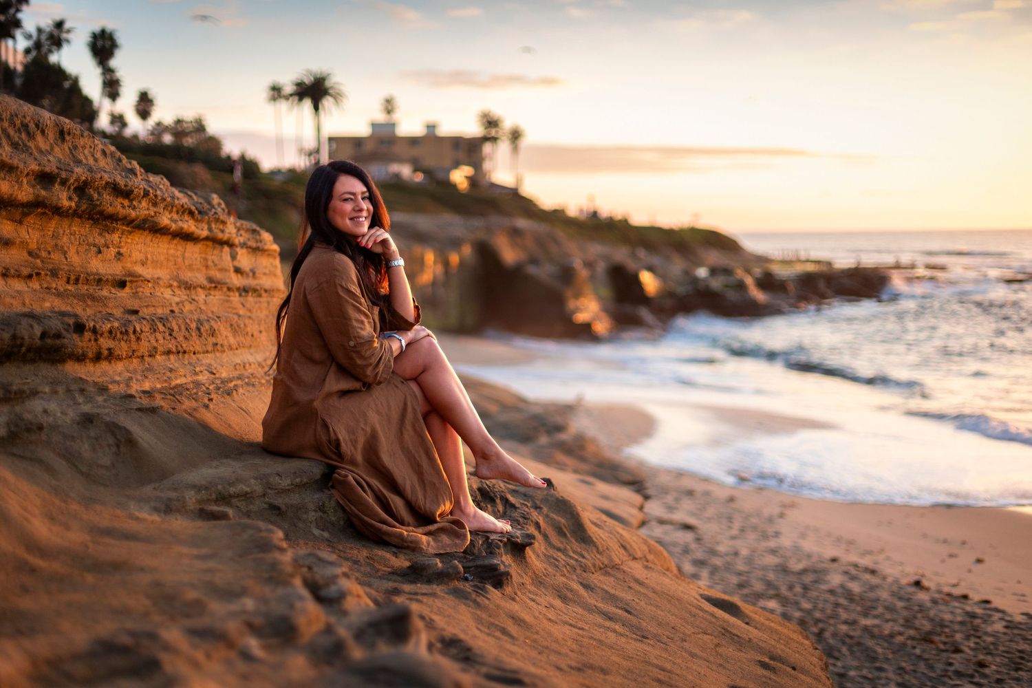 Woman in brown dress sitting on sandy cliffs at golden hour sunset with ocean waves and coastal cliffs behind her.