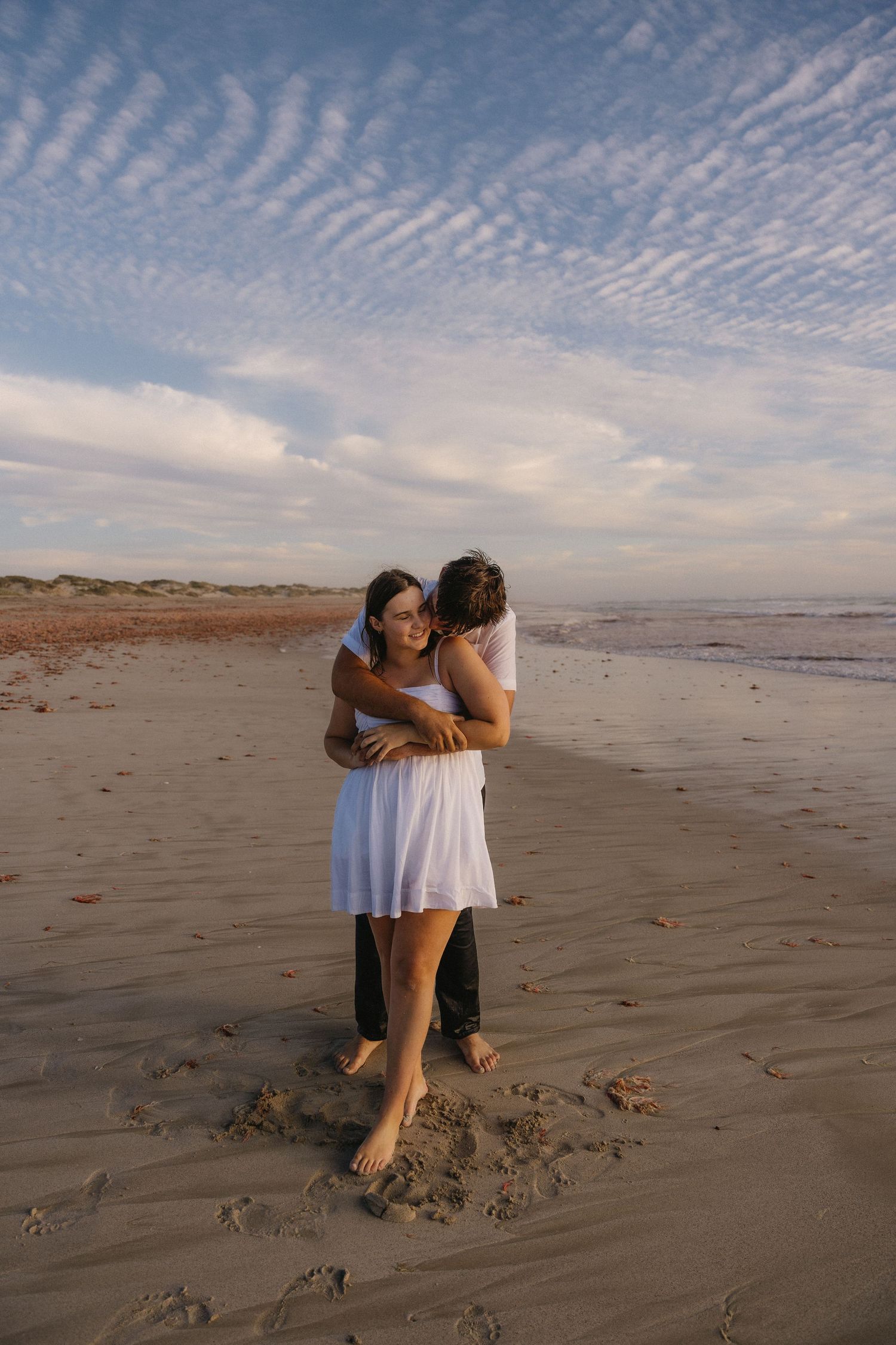 Two people playfully dance together on a sandy beach at sunset with rippled clouds in the sky above.