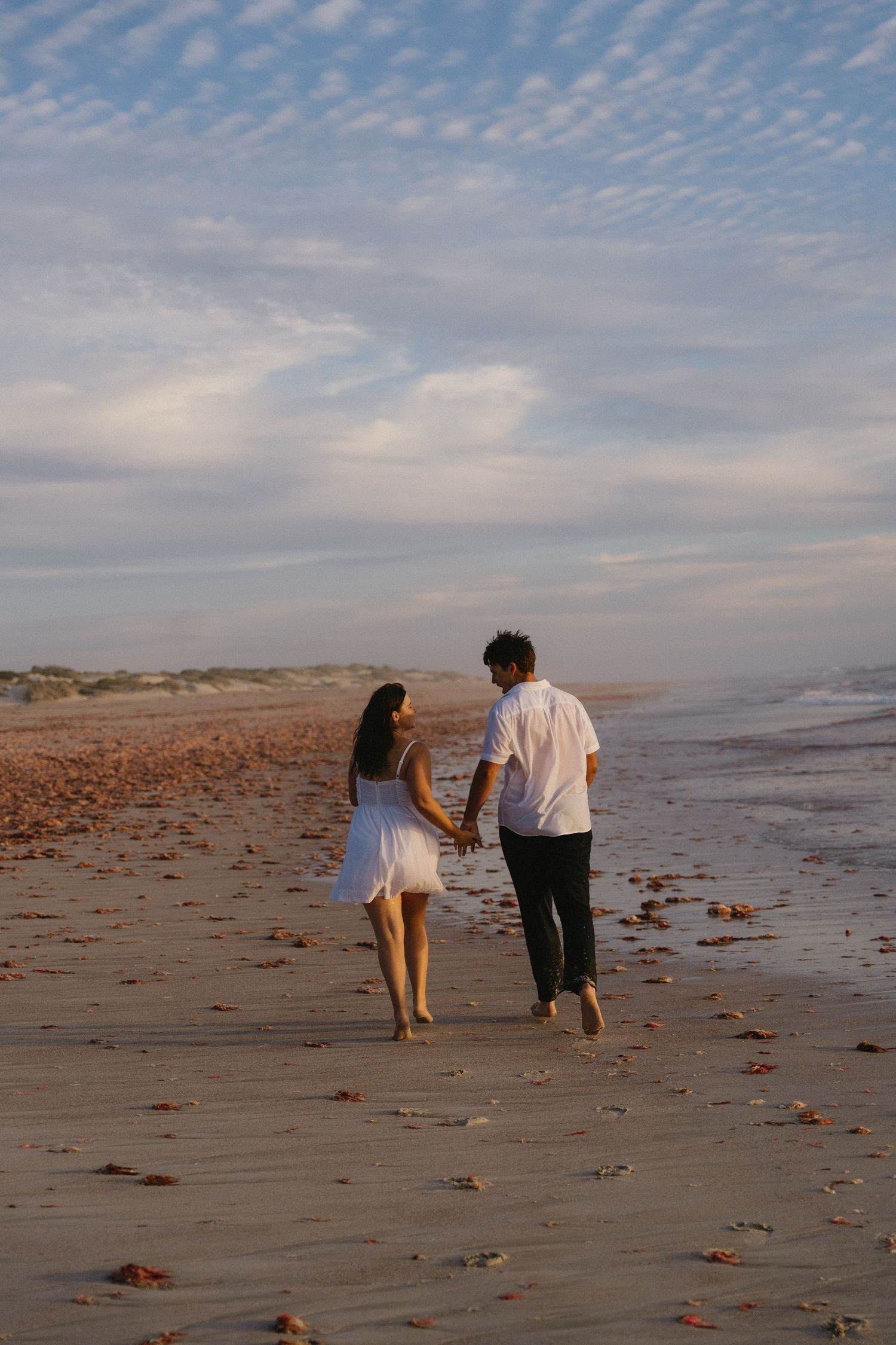 A couple strolls hand in hand along a serene beach at sunset with scattered seaweed on the sand.
