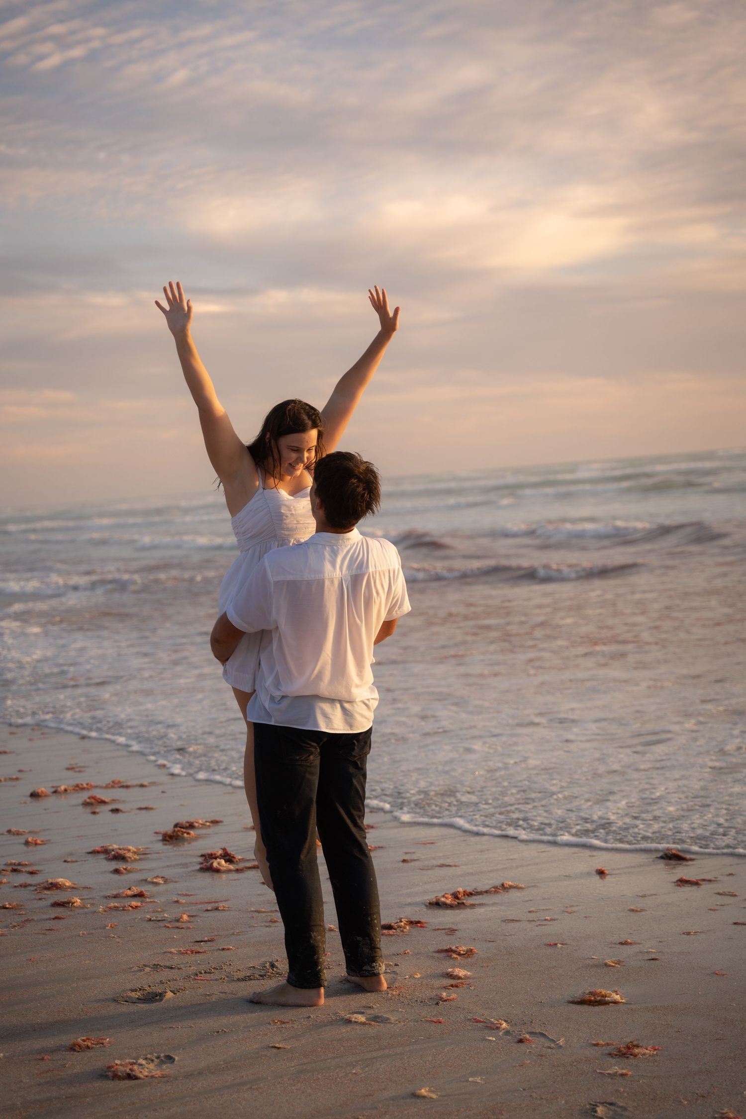 Two people in white shirts and jeans stand together on a beach at sunset with scattered autumn leaves on the sand.
