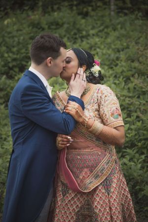 Couple sharing a romantic kiss outdoors, bride in traditional Indian lehenga with mehndi, groom in navy blue suit.