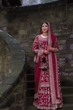 A bride in an ornate red and gold embroidered Pakistani bridal lehenga with dupatta, posing on stone steps.