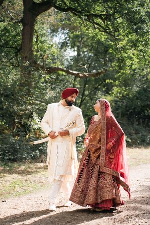 Sikh bride and groom in traditional attire walk together on a sunlit forest path, groom holding a ceremonial kirpan.