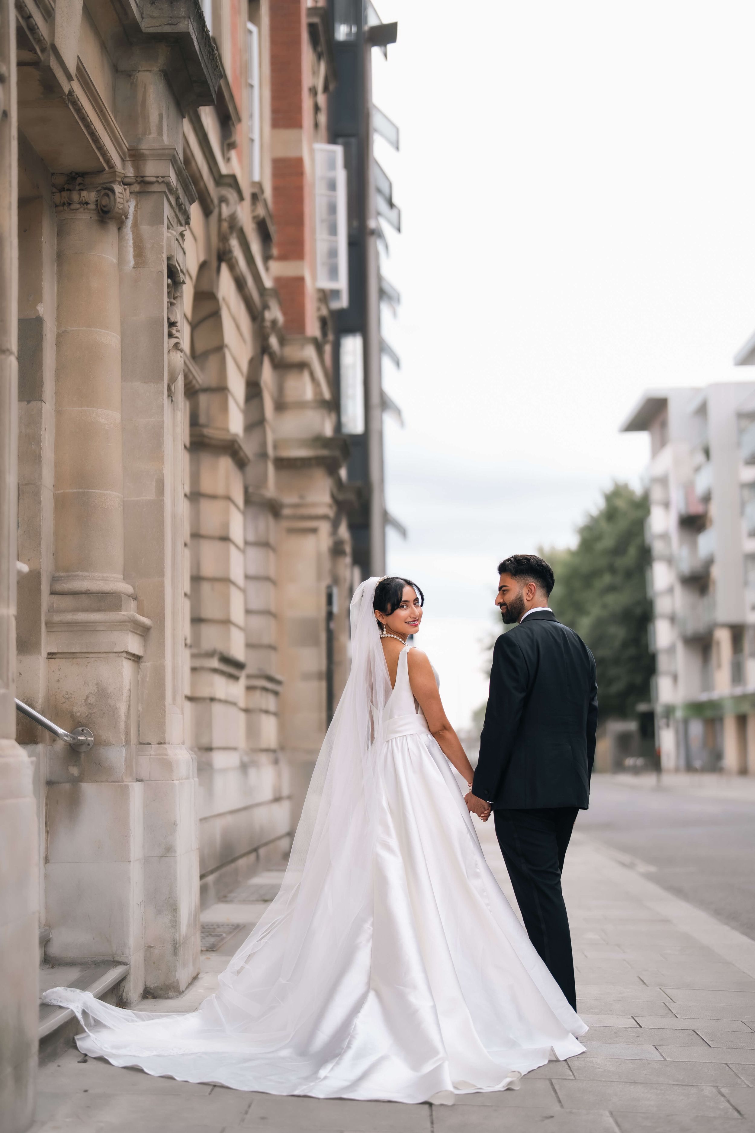 Bride in white gown and groom in black suit hold hands on a city street beside a classic stone building.