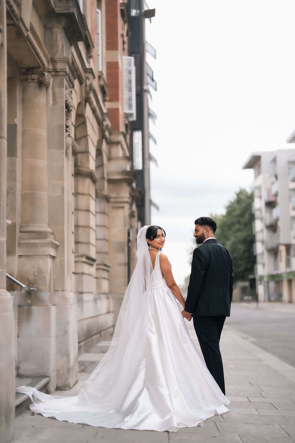 Bride in white gown and groom in black suit hold hands on a city street beside a classic stone building.
