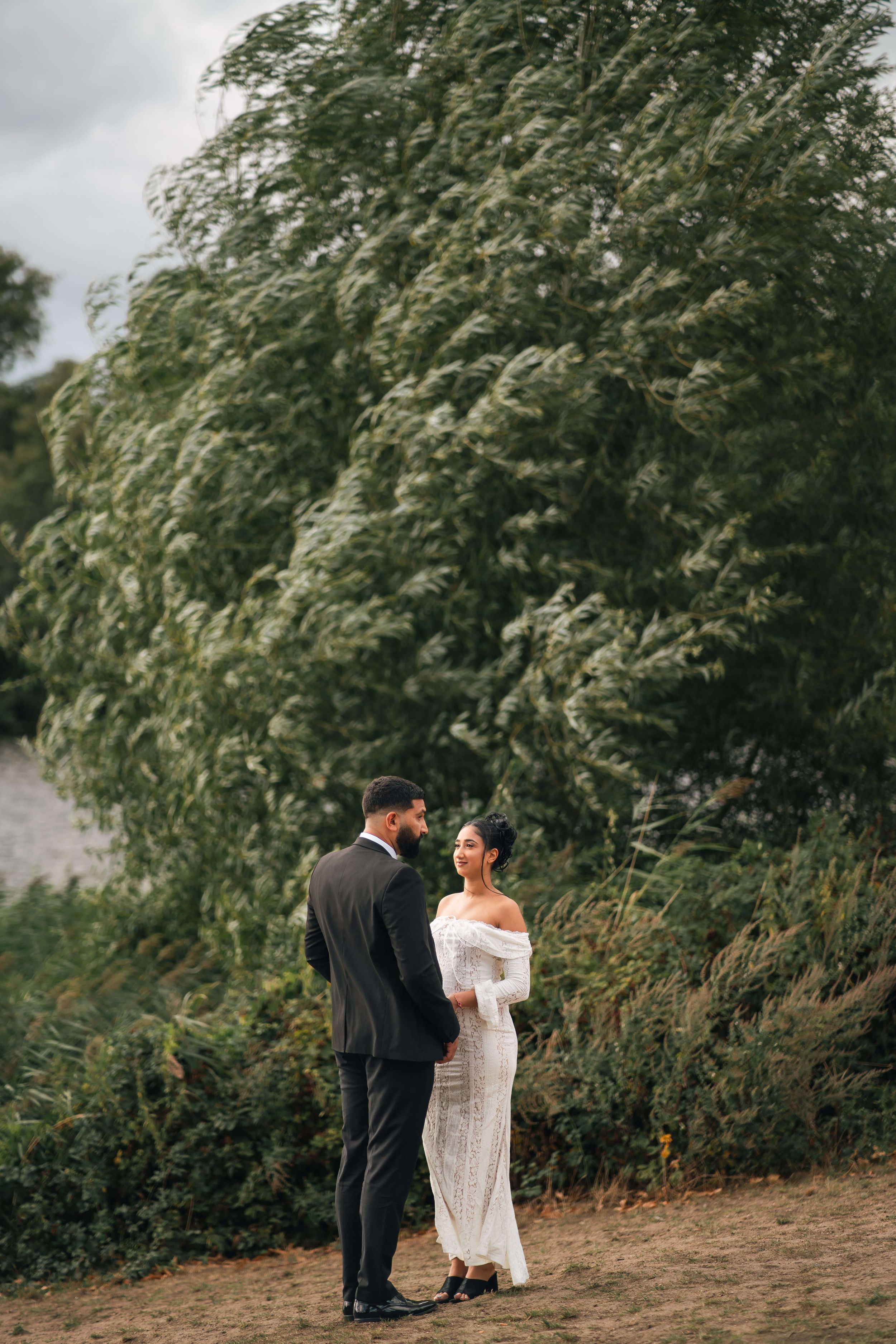 A bride in a white off-shoulder gown and groom in black tuxedo stand together outdoors near lush green trees.