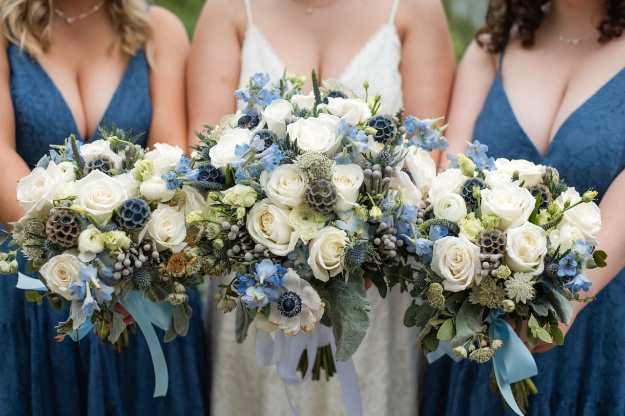 Wedding bouquets featuring white roses and blue flowers held by bridesmaids in navy dresses.