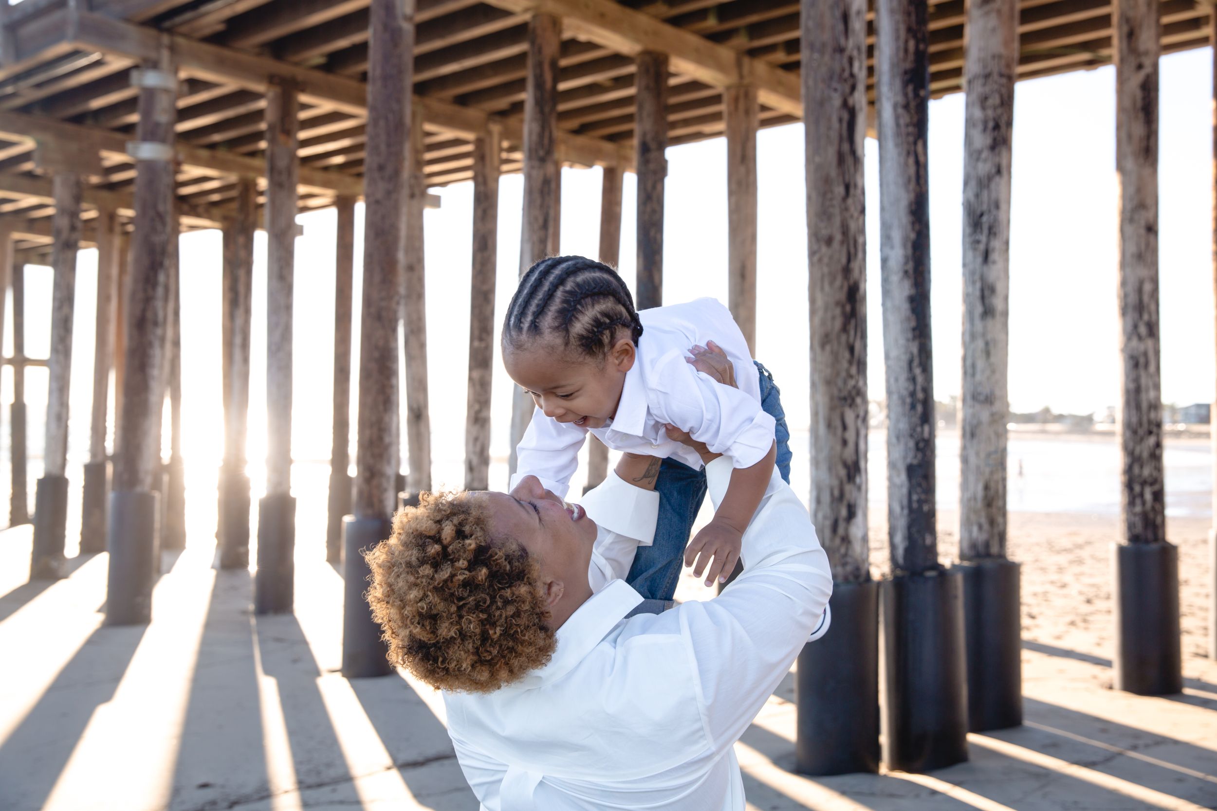 A loving parent lifts their child playfully under wooden pier posts at sunset creating long dramatic shadows.