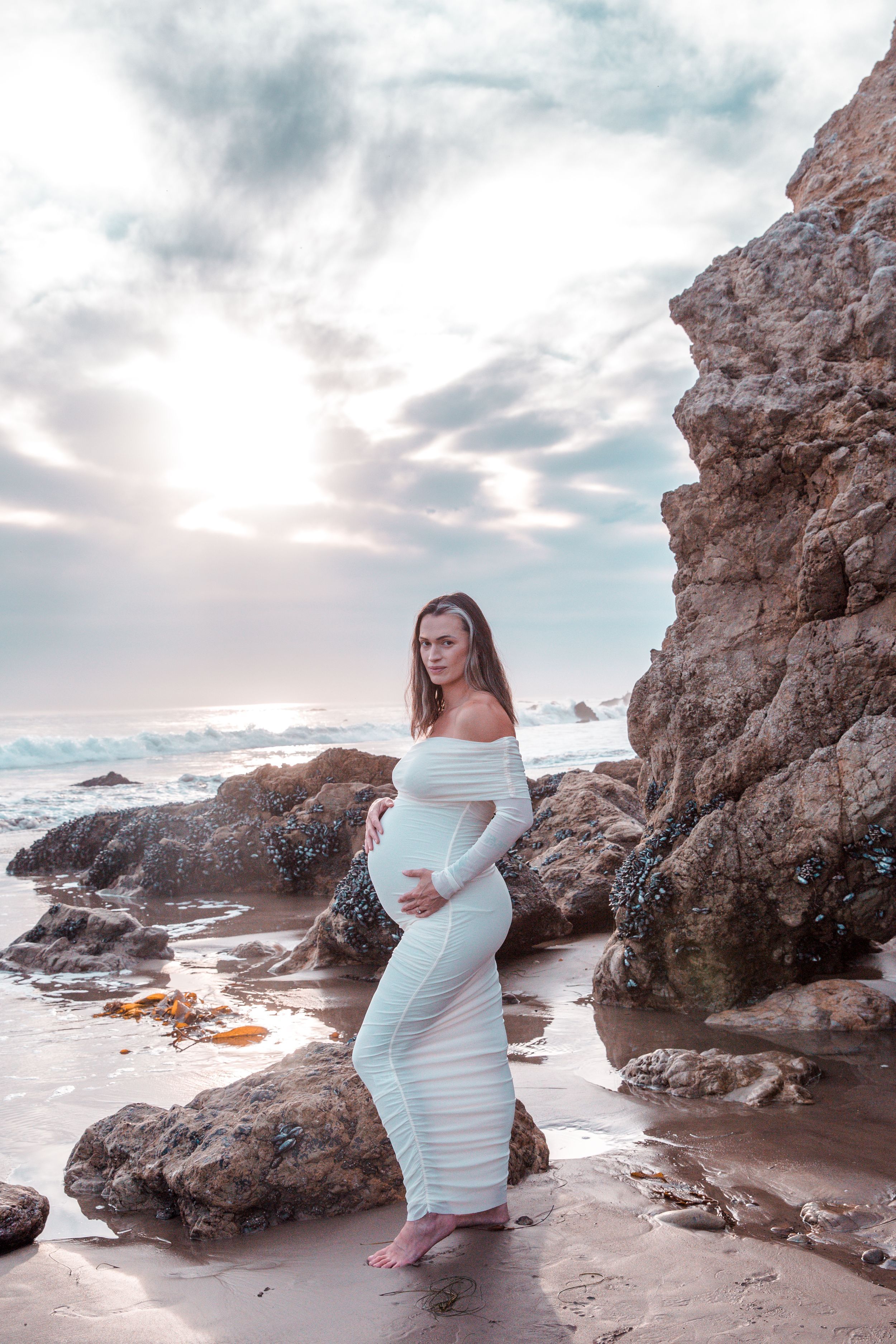 An expectant woman in a white flowing dress poses on a beach at sunset among rocky coastal formations.