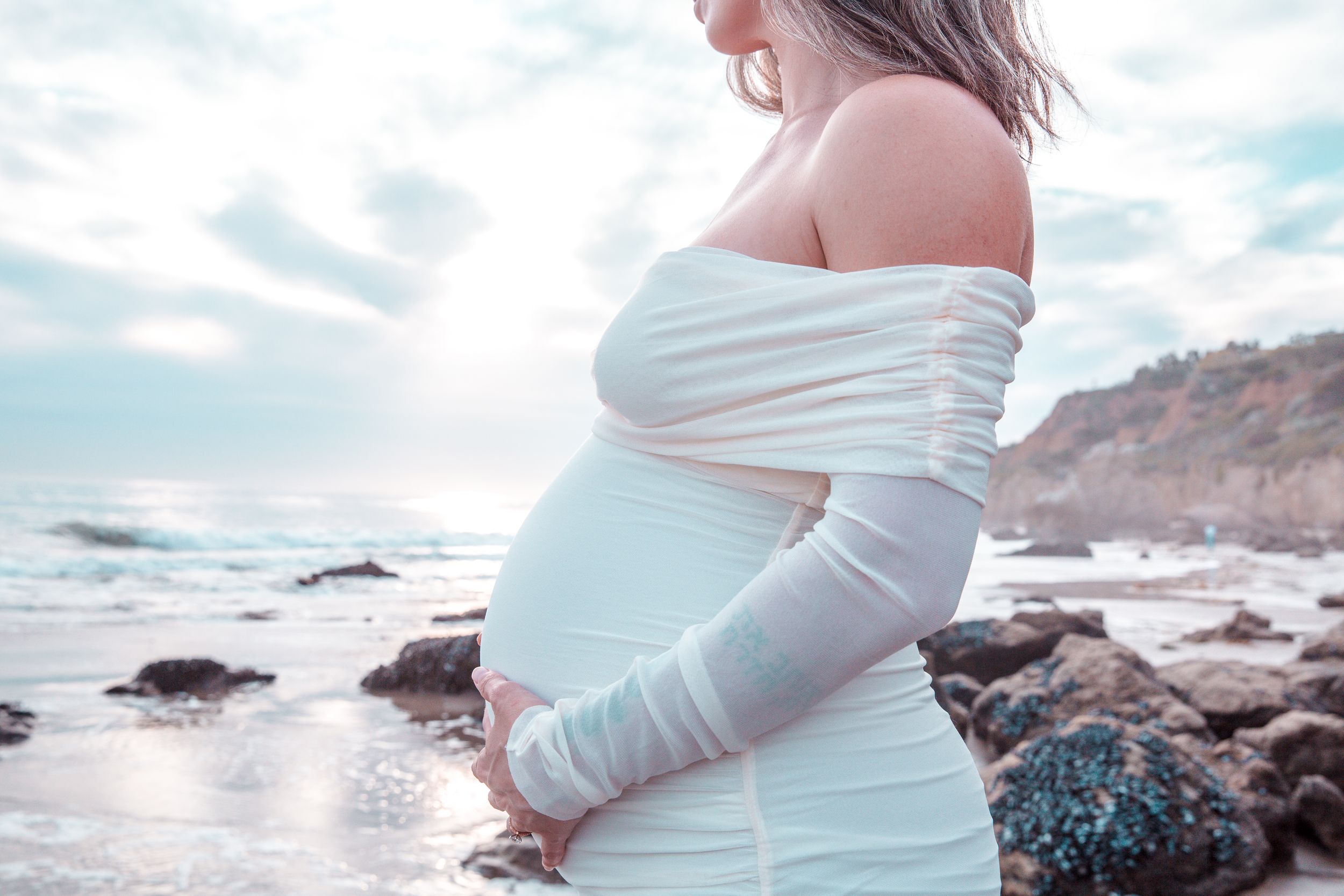 A pregnant woman in a white dress stands on a rocky beach at sunset, cradling her baby bump.