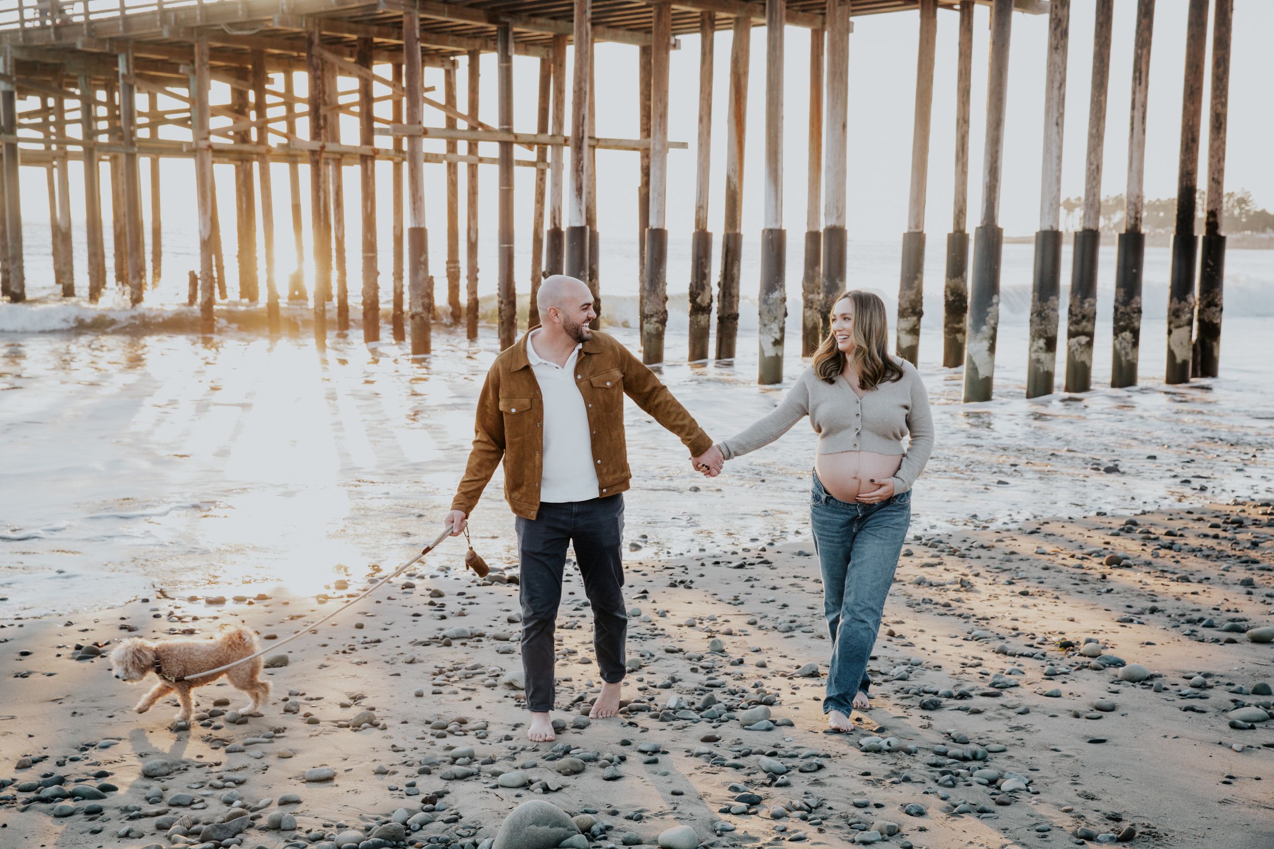 A pair strolls hand in hand on the beach beneath a wooden pier during golden sunset hour at low tide.