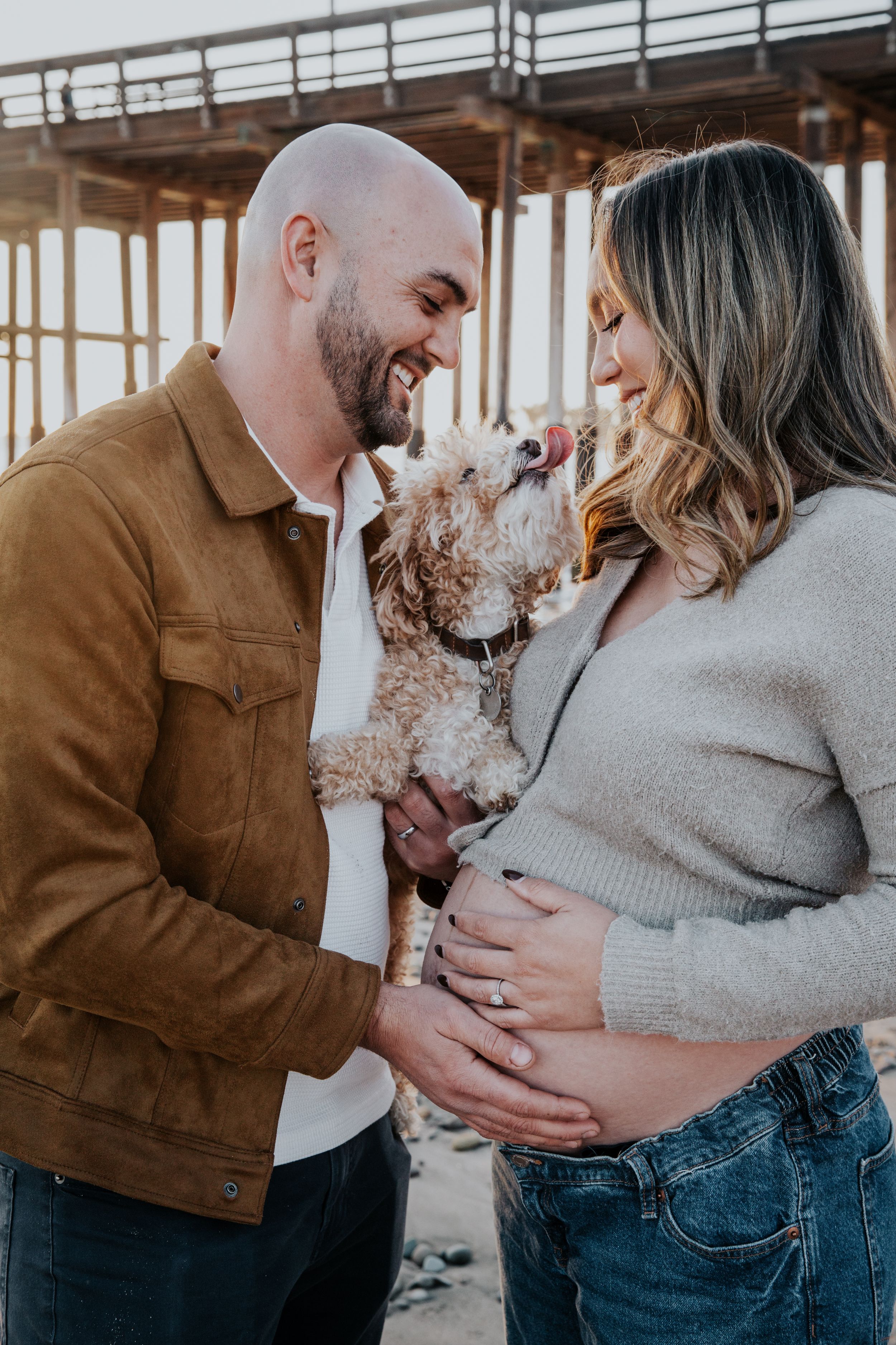 Expecting couple shares joyful moment with their small curly-haired dog under pier.