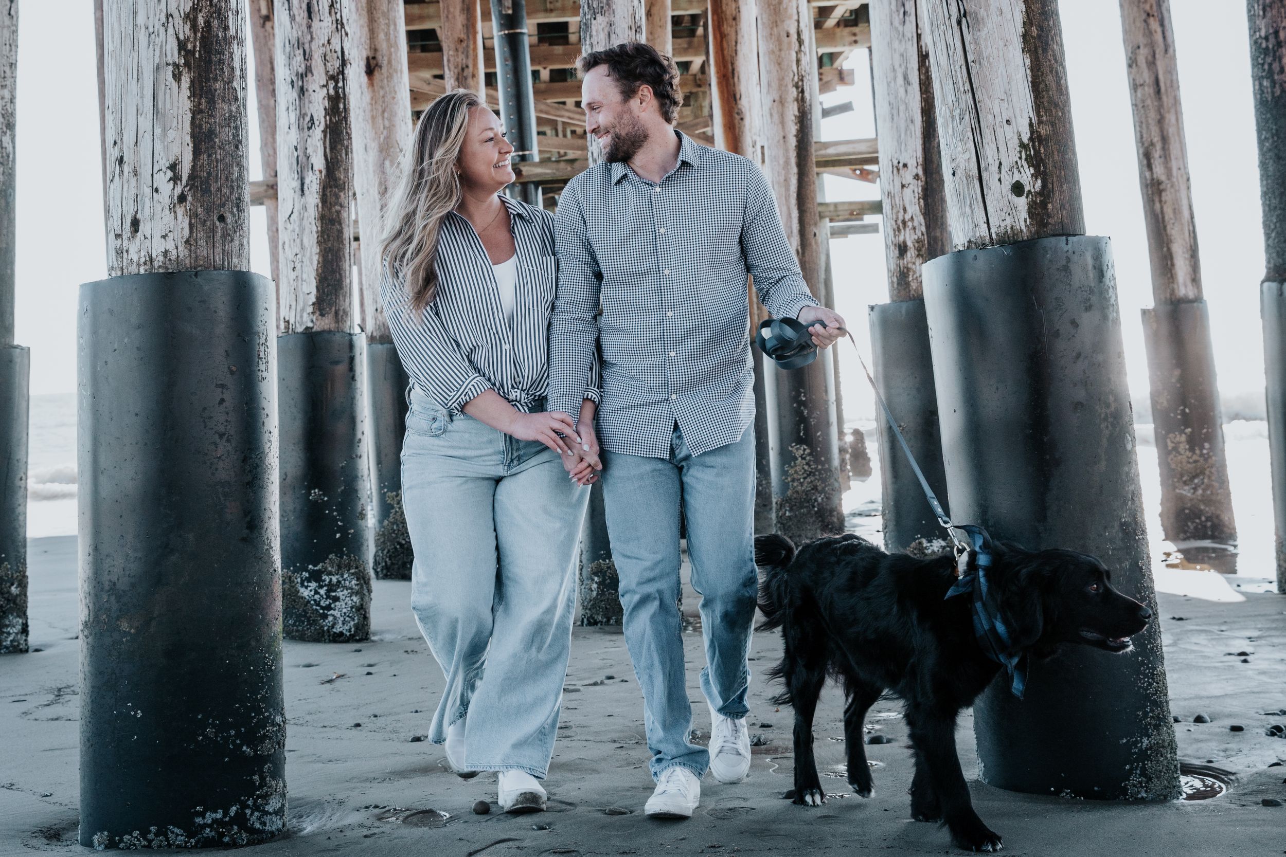 A couple walks their black dog under a pier on the beach while wearing matching casual outfits.