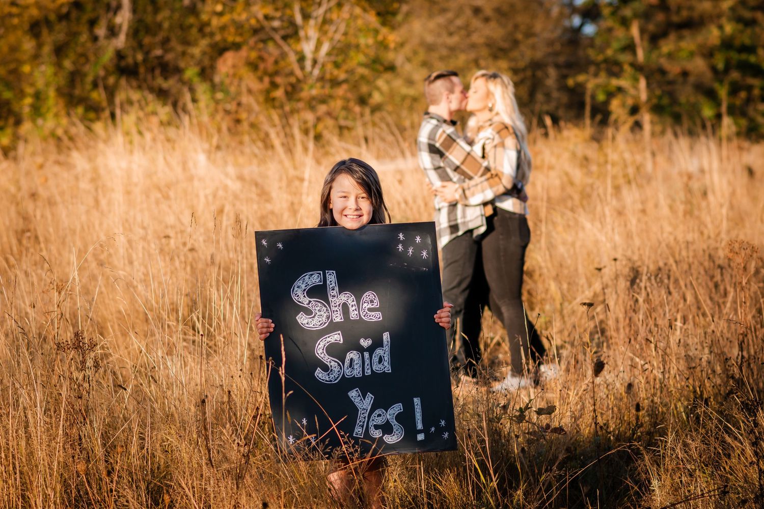 Someone holds a chalkboard sign in a golden field with their dog beside them.