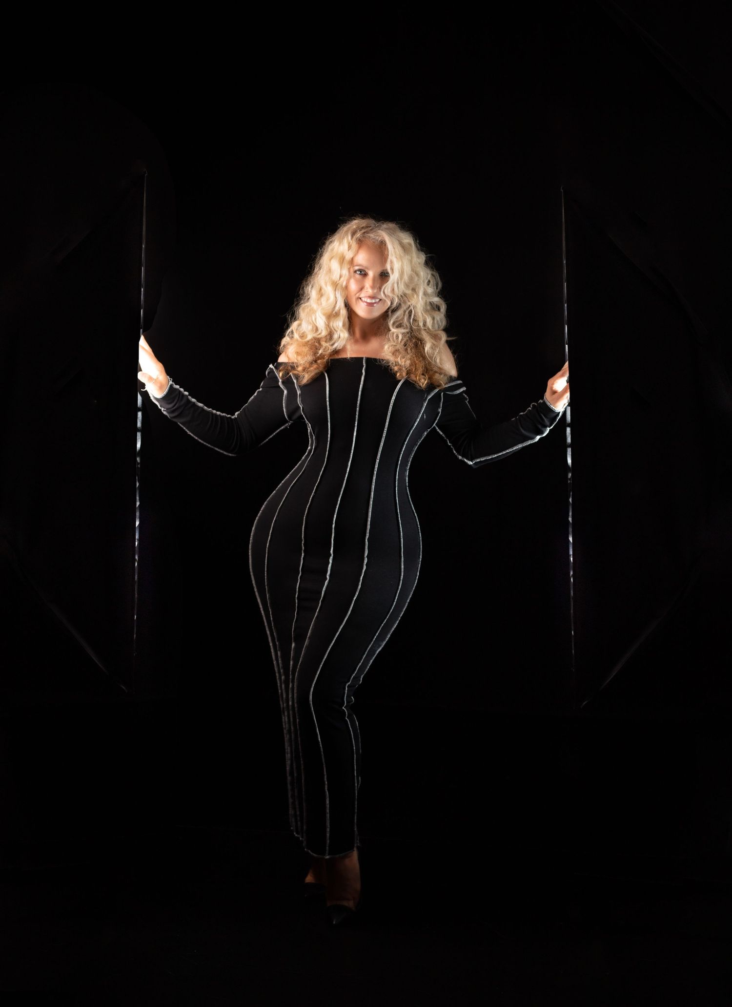 A striking woman with an hourglass figure in a black striped dress poses in dramatic light against a dark background.
