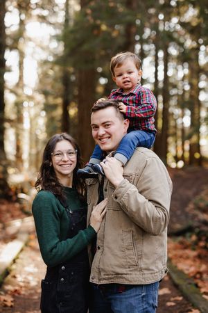 A mother and father holding hands with their toddler while walking down a sun-dappled wooded forest trail in Snohomish.