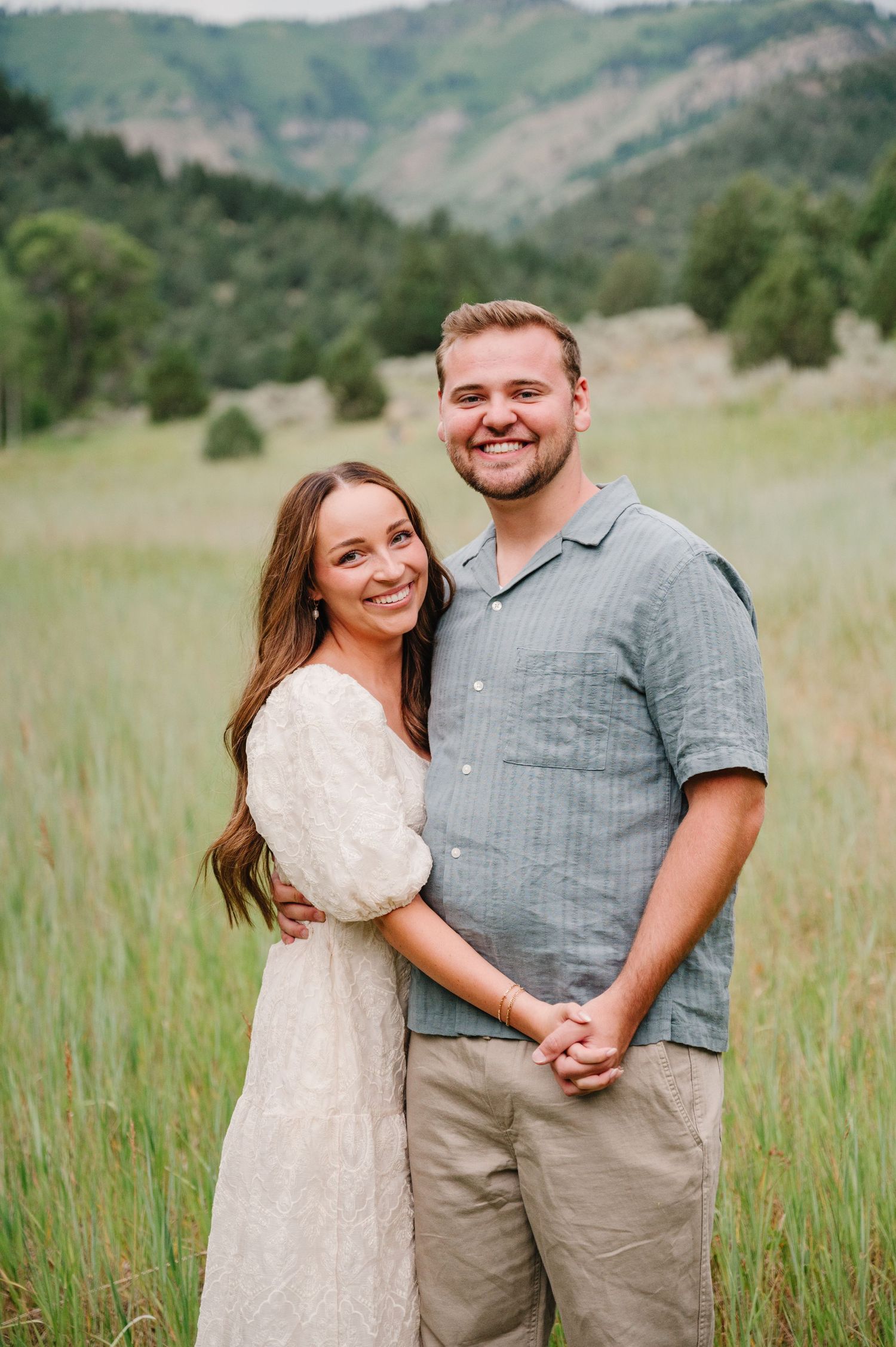 A couple poses together in a field with mountain scenery, wearing casual summer clothing on a sunny afternoon.