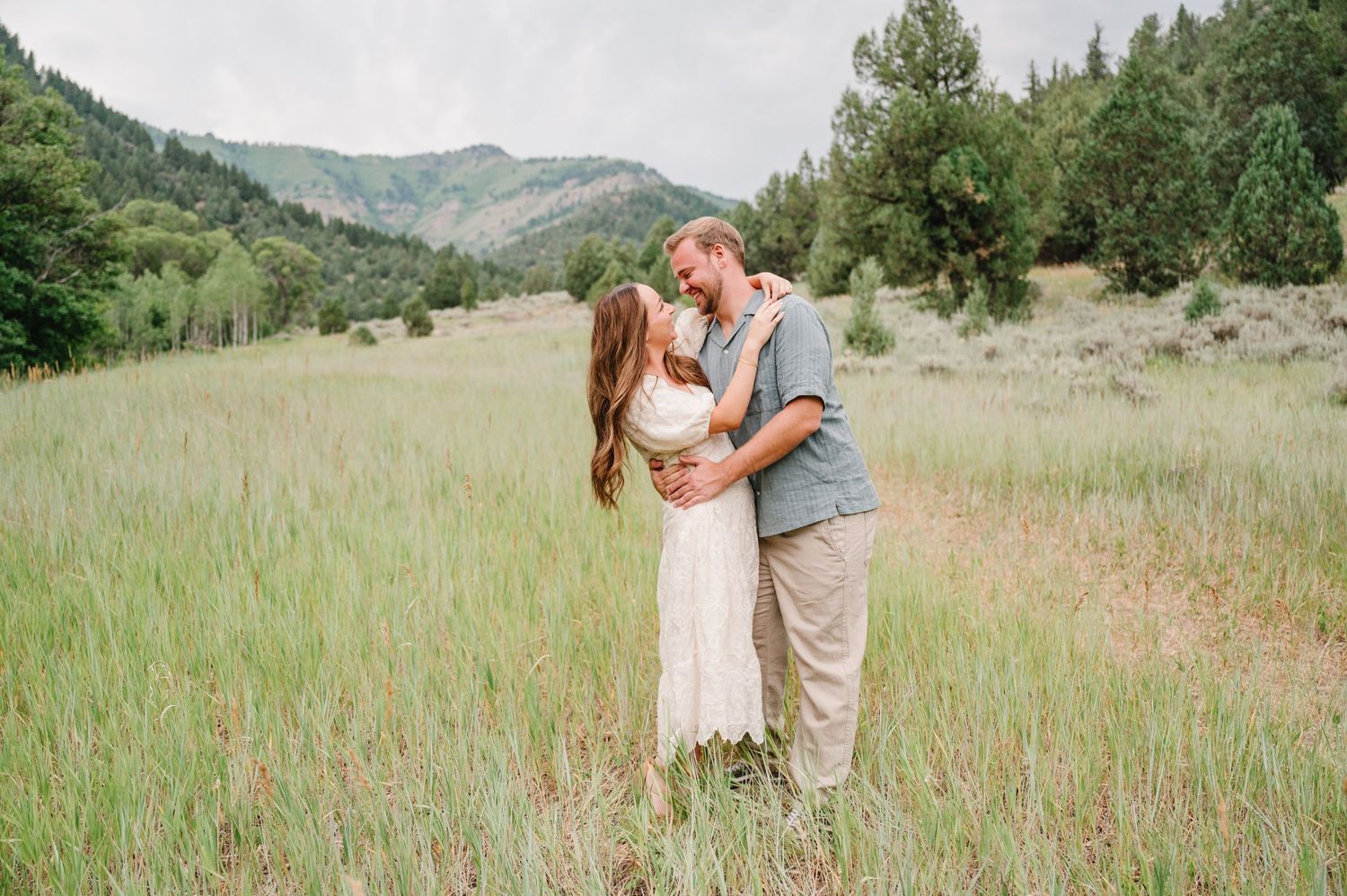 A couple embraces in a romantic moment while standing in a scenic mountain meadow filled with tall grass.