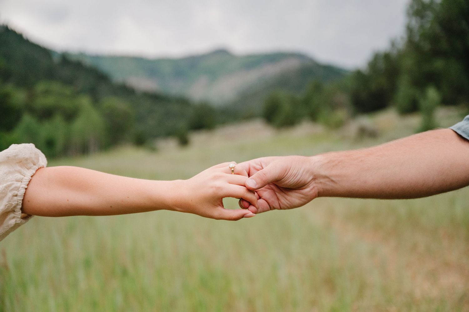 Two hands reaching out to hold each other in a tender moment against a backdrop of green rolling hills and mountains.