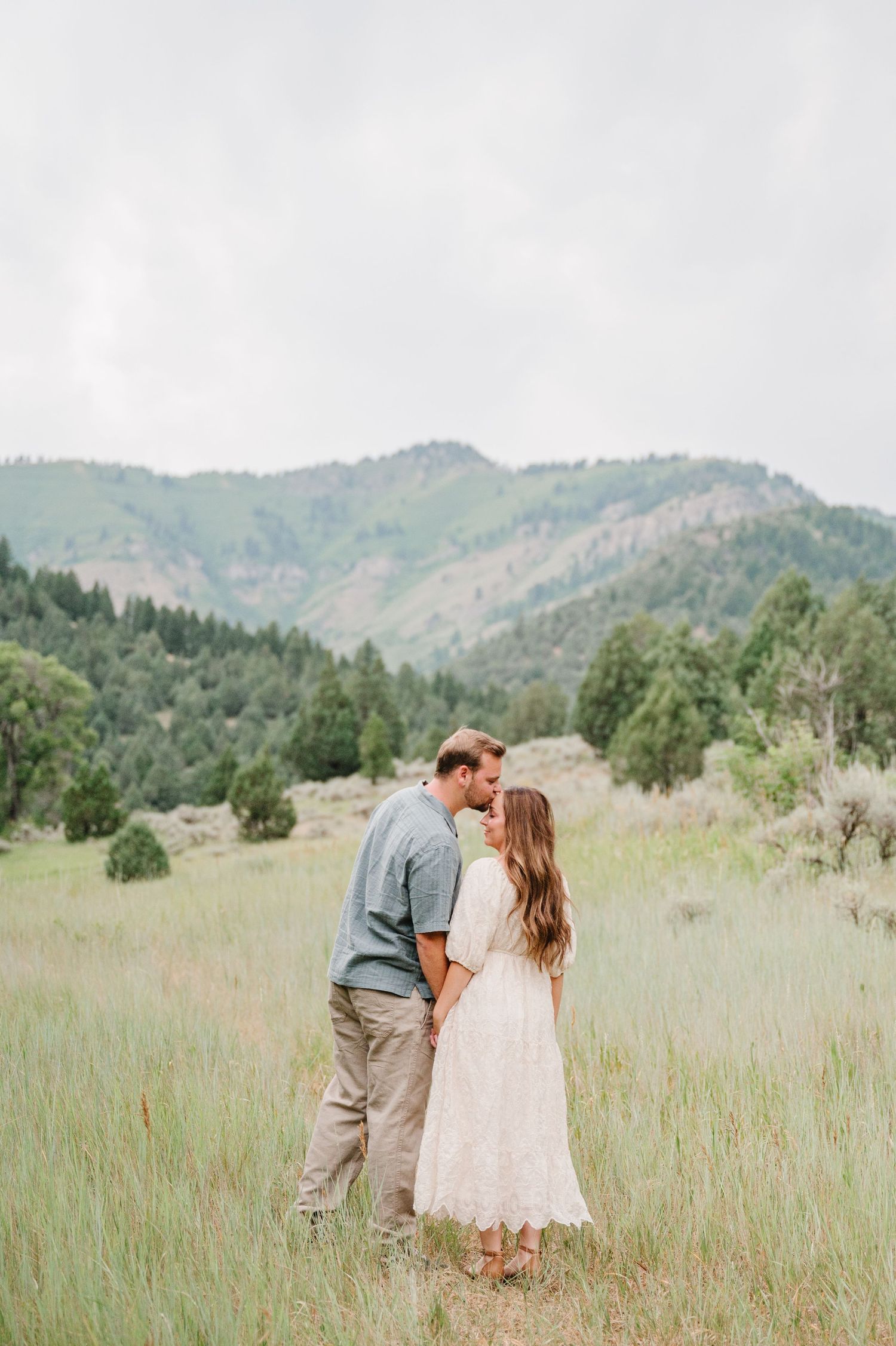 A couple shares a romantic moment in a meadow with mountains and trees in the background.