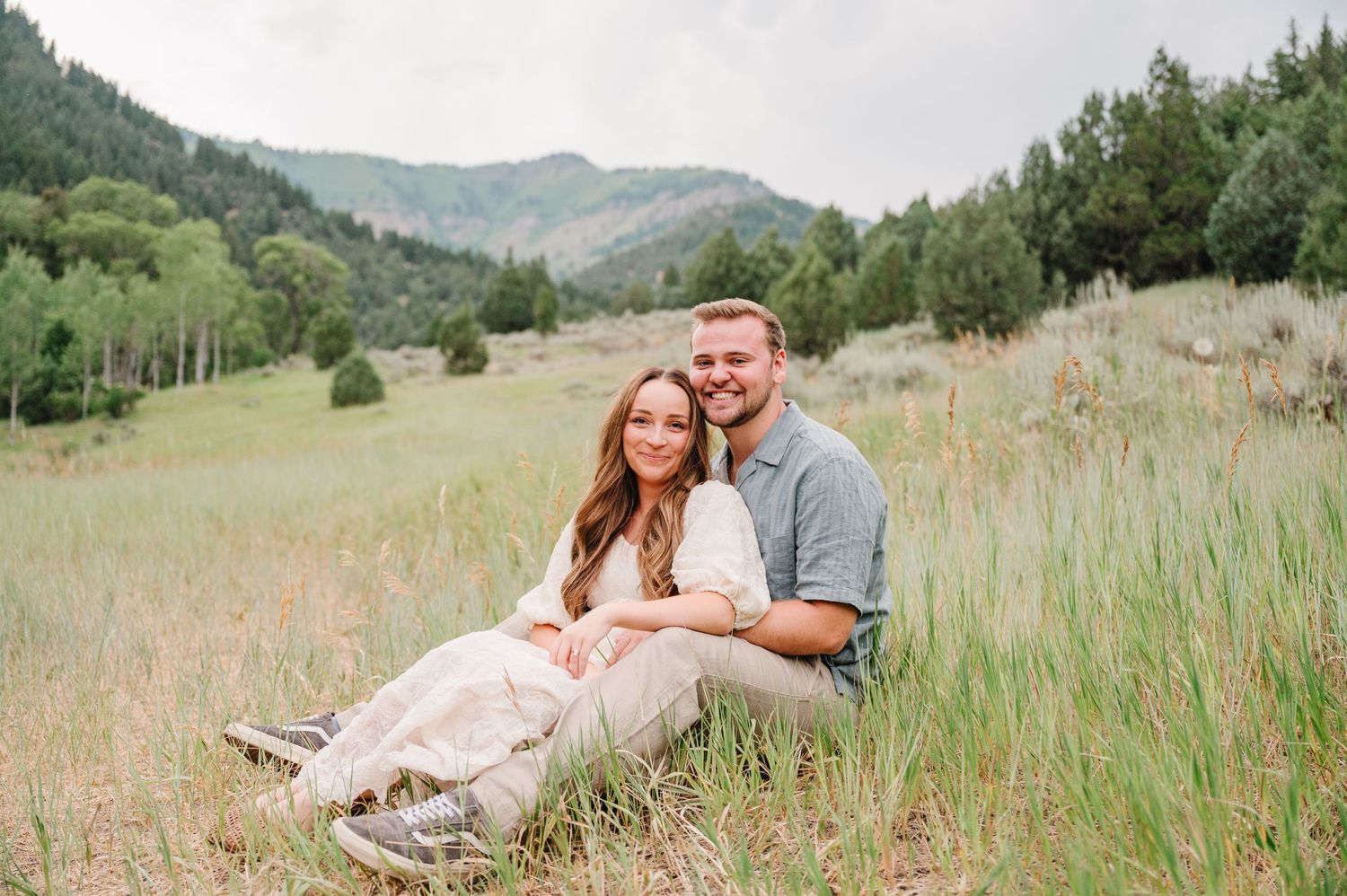 A couple sits together in a meadow surrounded by rolling green hills and tall grass on a summer afternoon.