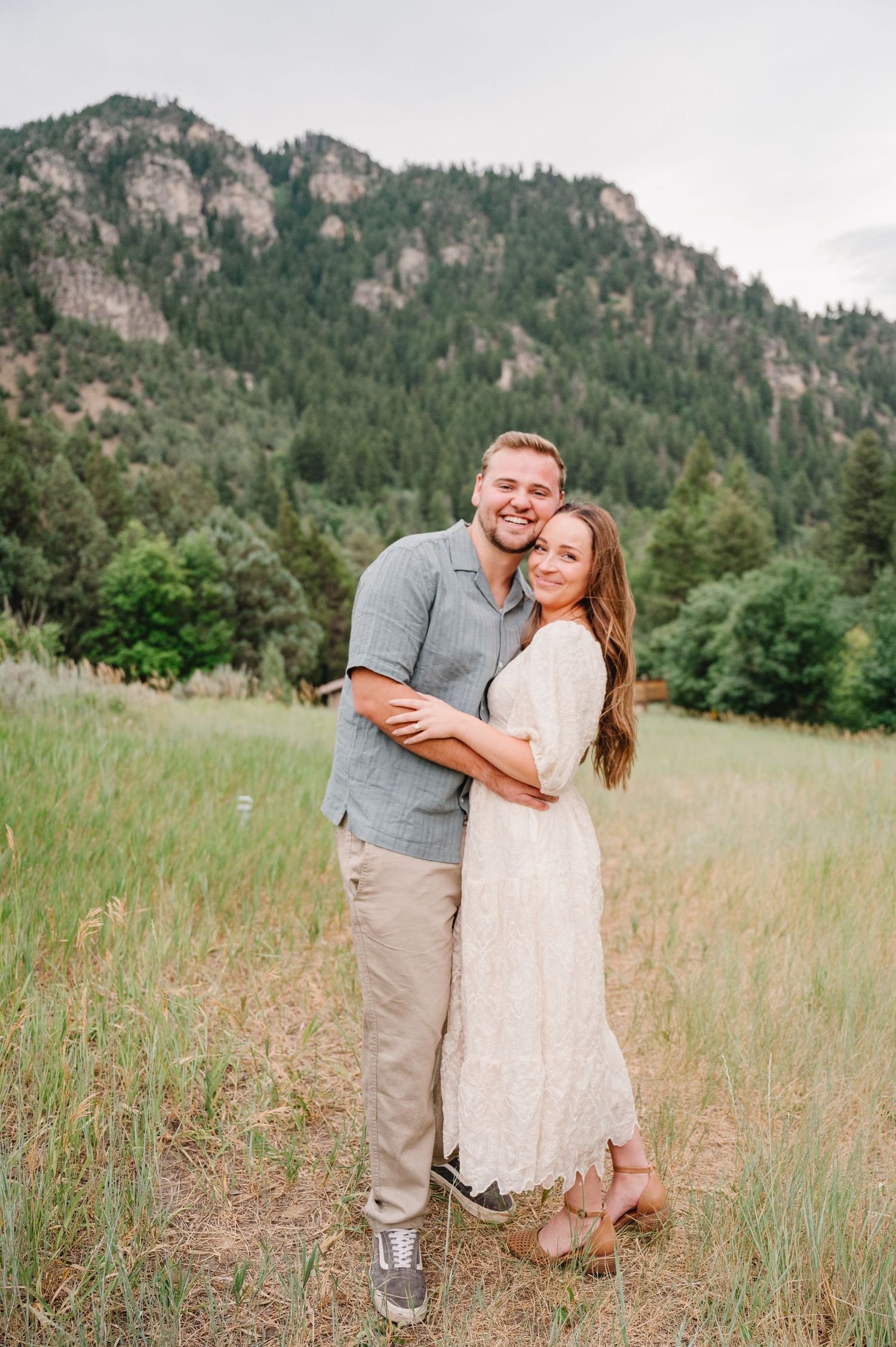 A couple embraces in a scenic mountain meadow with tall rocky cliffs and evergreen forest in the background.