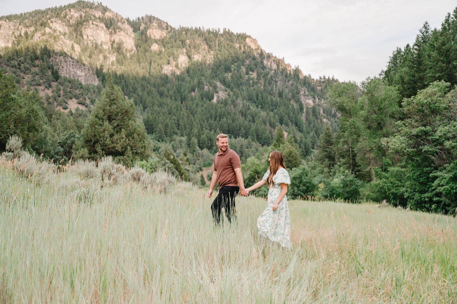 Couple walking together through tall grass on mountain meadow with rocky cliffs and evergreen forest in background.