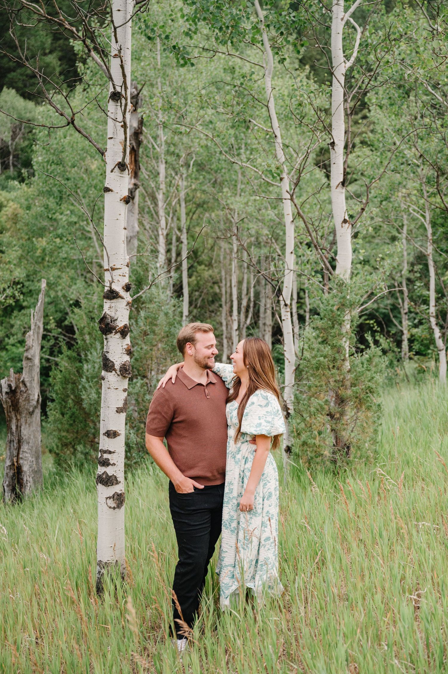 A couple share a tender moment among tall aspen trees in a lush green mountain meadow.