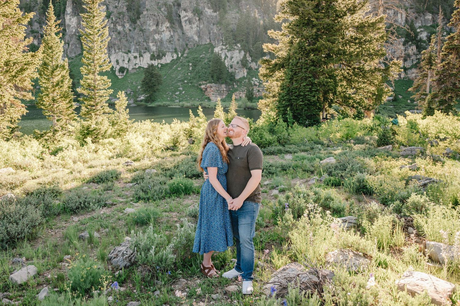 Couple embracing in a lush mountain meadow surrounded by tall pine trees and rocky cliffs during golden hour.