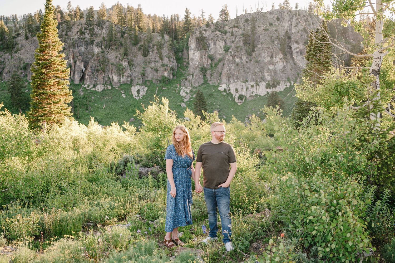 Couple standing in lush green mountain meadow surrounded by trees and rocky cliffs in golden hour sunlight.