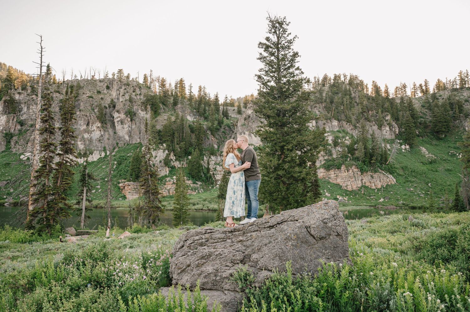 Couple embracing on a large rock surrounded by wildflowers with a mountain lake and pine forest backdrop.