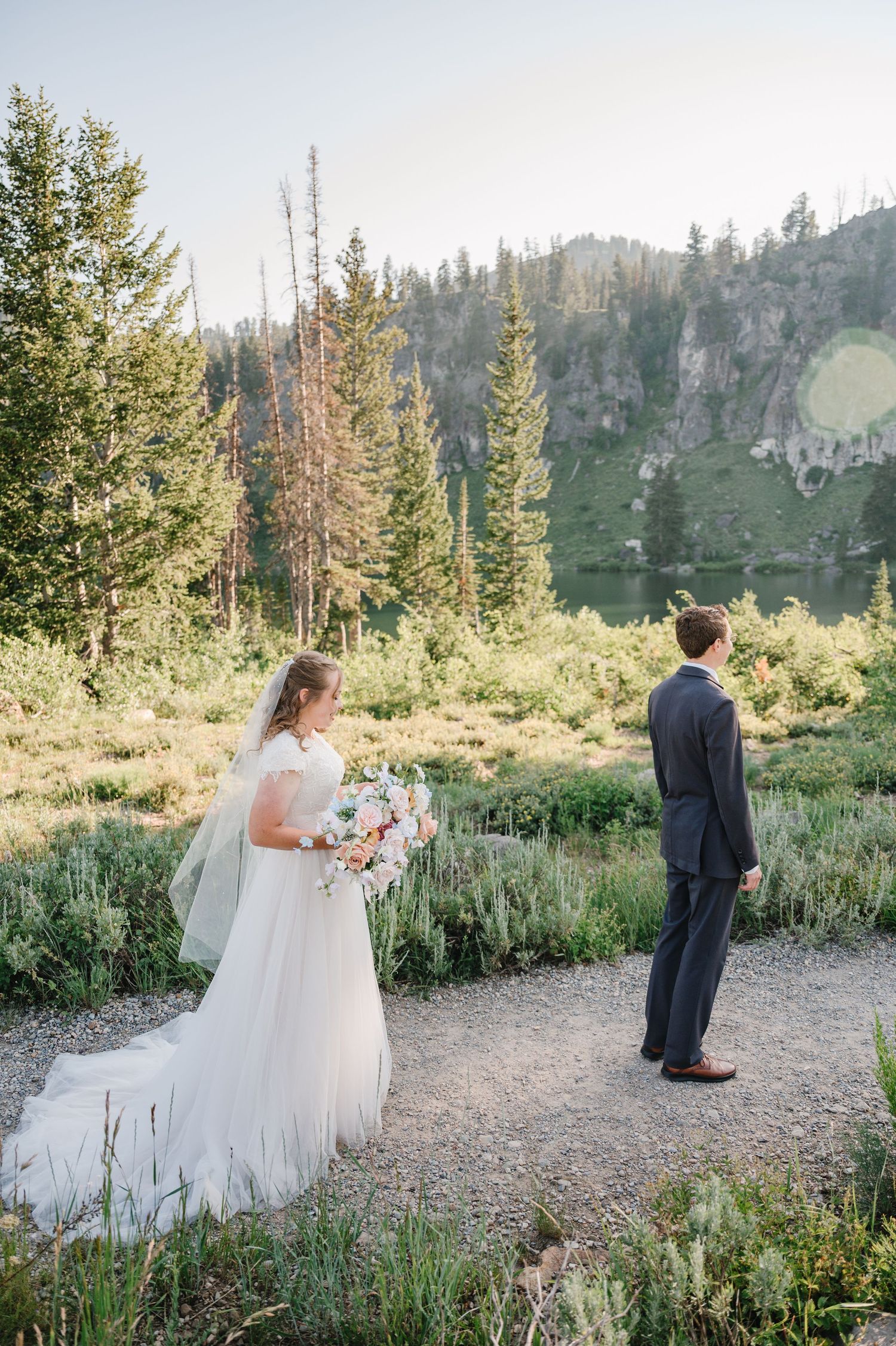 A couple sharing a first look moment on a mountain trail surrounded by pine trees and wildflowers.