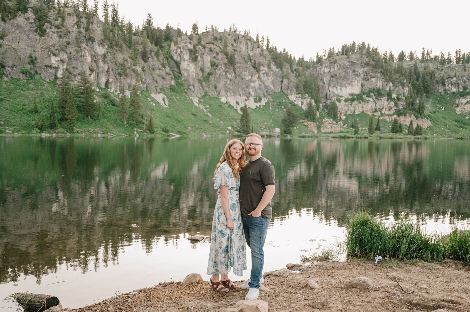 Couple smiling together by a serene mountain lake with rocky cliffs and pine trees reflected in calm water.