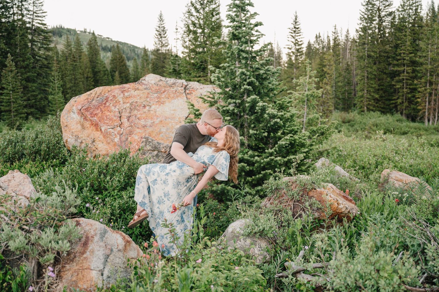 Couple sharing a romantic dip kiss among lush green shrubs and boulders in a mountain forest setting.