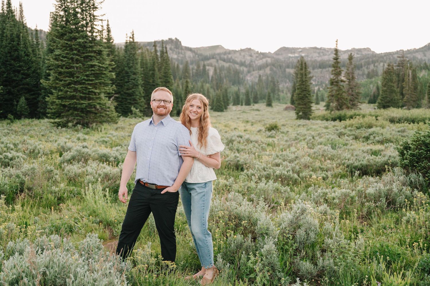 Couple smiling together in a mountain meadow surrounded by evergreen trees and scenic peaks.