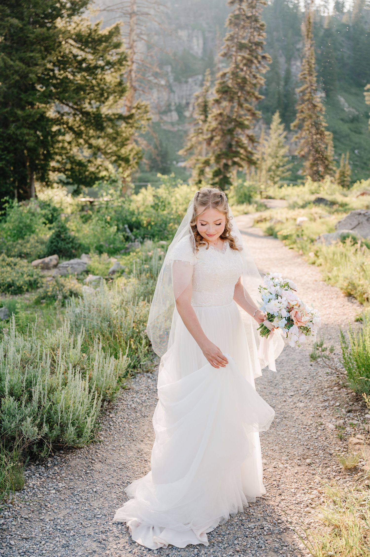 A bride walks along a mountain trail holding a bouquet of flowers.