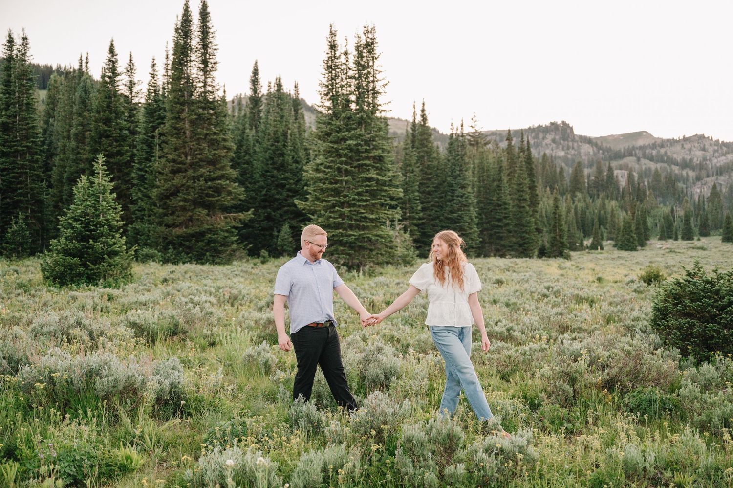 Couple holding hands walking through a lush green mountain meadow surrounded by tall evergreen trees.