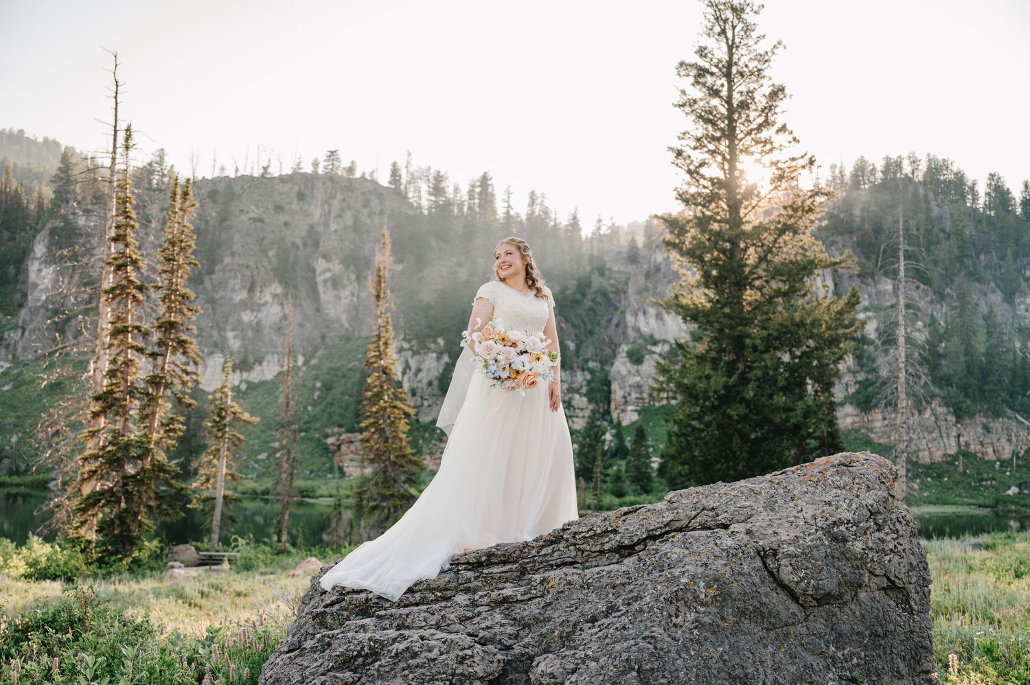 A bride in a flowing white dress stands atop a rocky outcrop with mountain views.