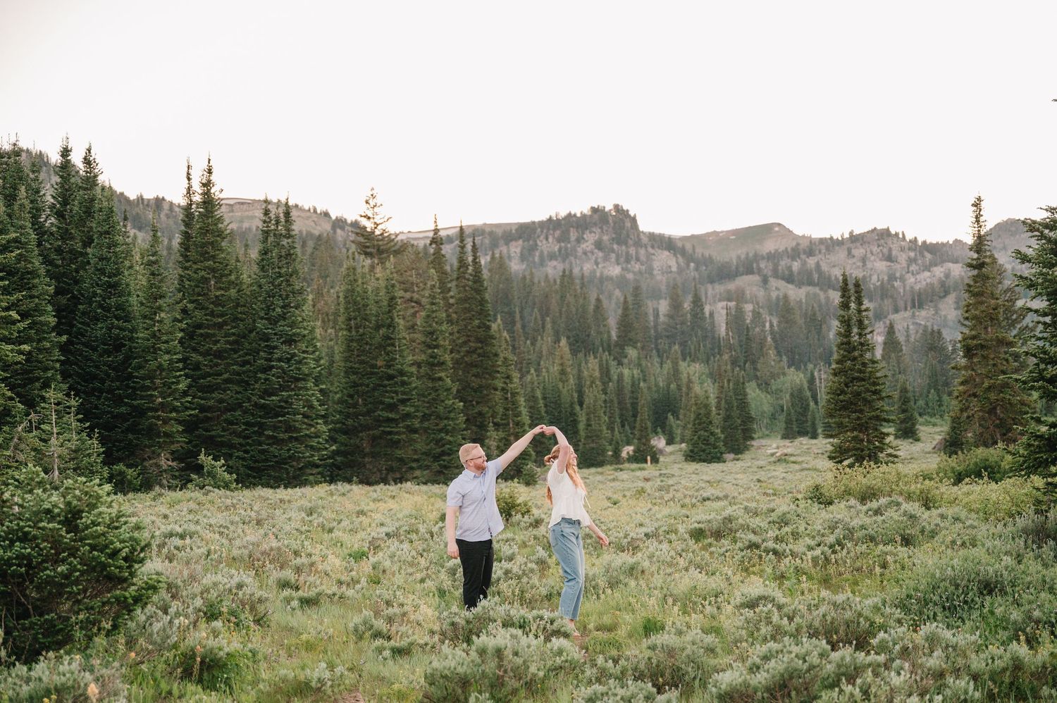 Couple dancing in a scenic mountain meadow surrounded by evergreen trees and rolling hills at dusk.