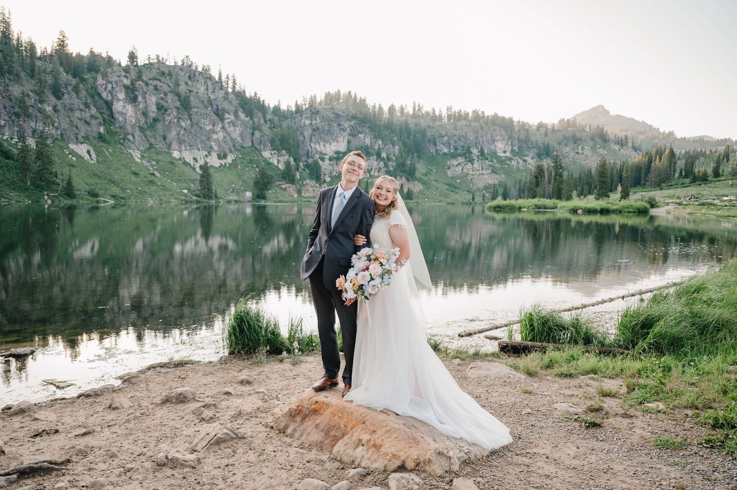 A romantic wedding photo of a couple by a serene mountain lake surrounded by evergreen trees.