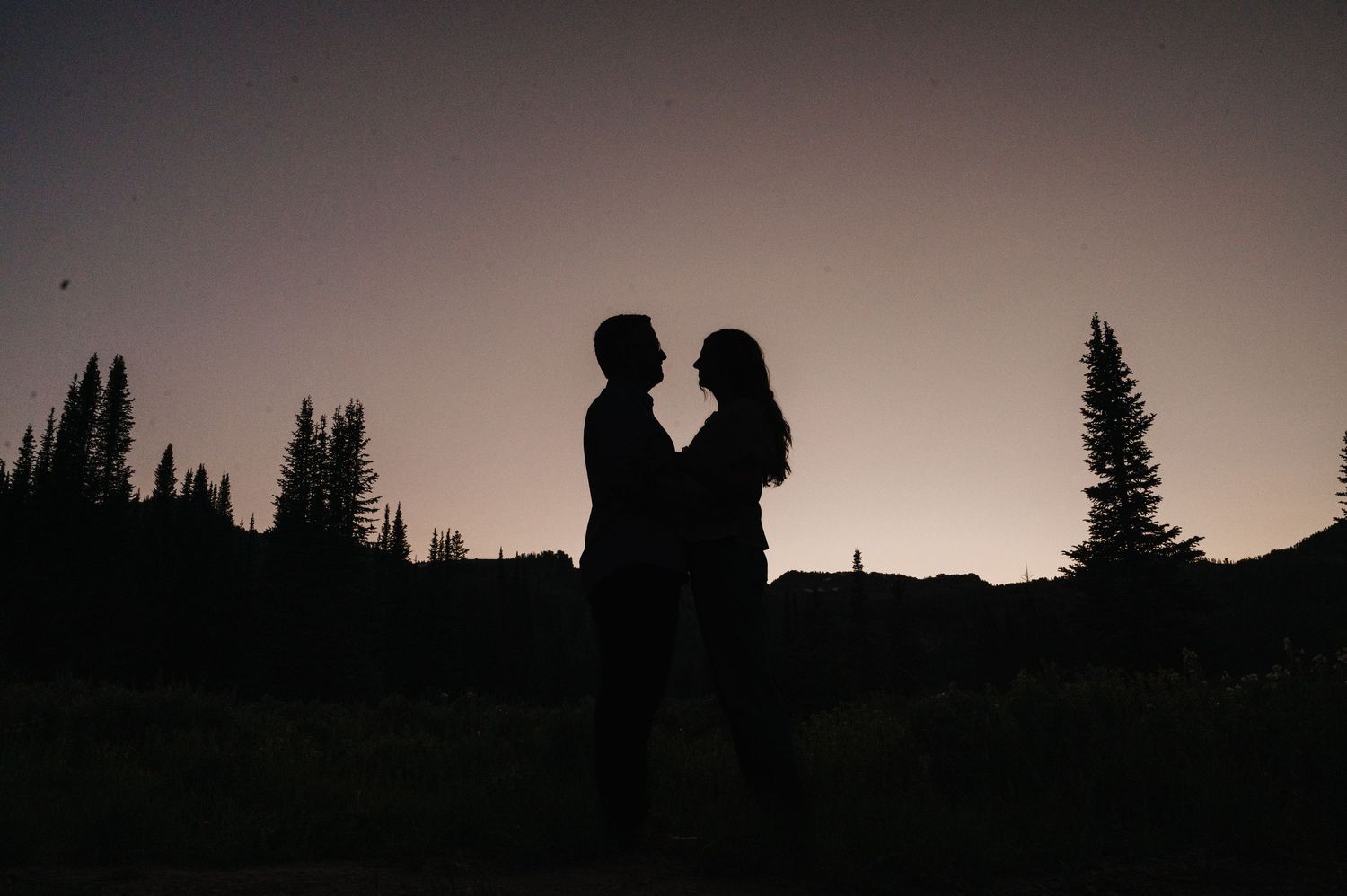 Silhouette of a romantic couple facing each other at dusk with pine trees and twilight sky backdrop.