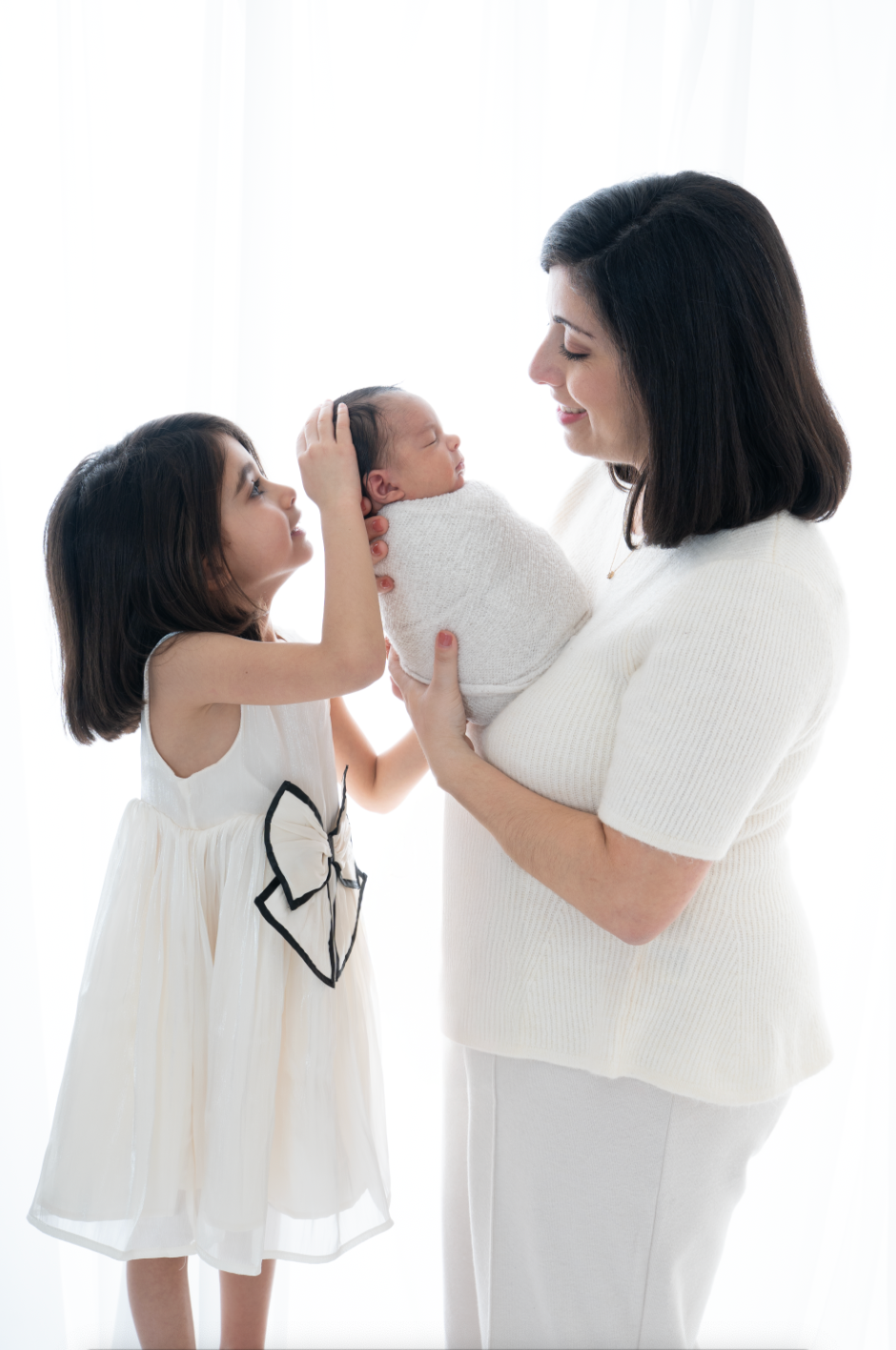 Mother in white holds swaddled newborn while young daughter in white dress gently touches baby's head in bright studio.