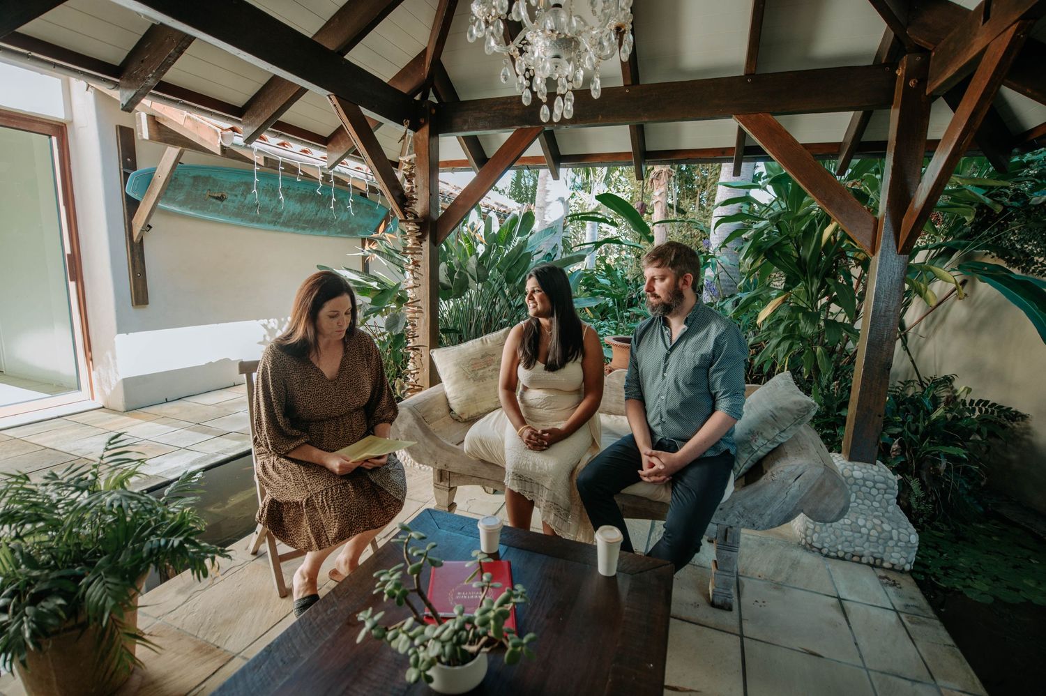 Three people sit together under a wooden pergola surrounded by tropical plants.