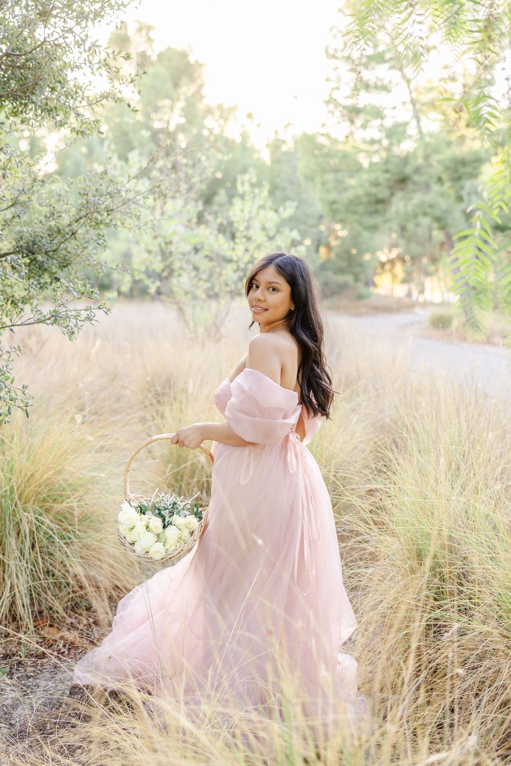 A person in a flowing pink off-shoulder dress holds a white flower basket while standing in tall grass during golden hour.