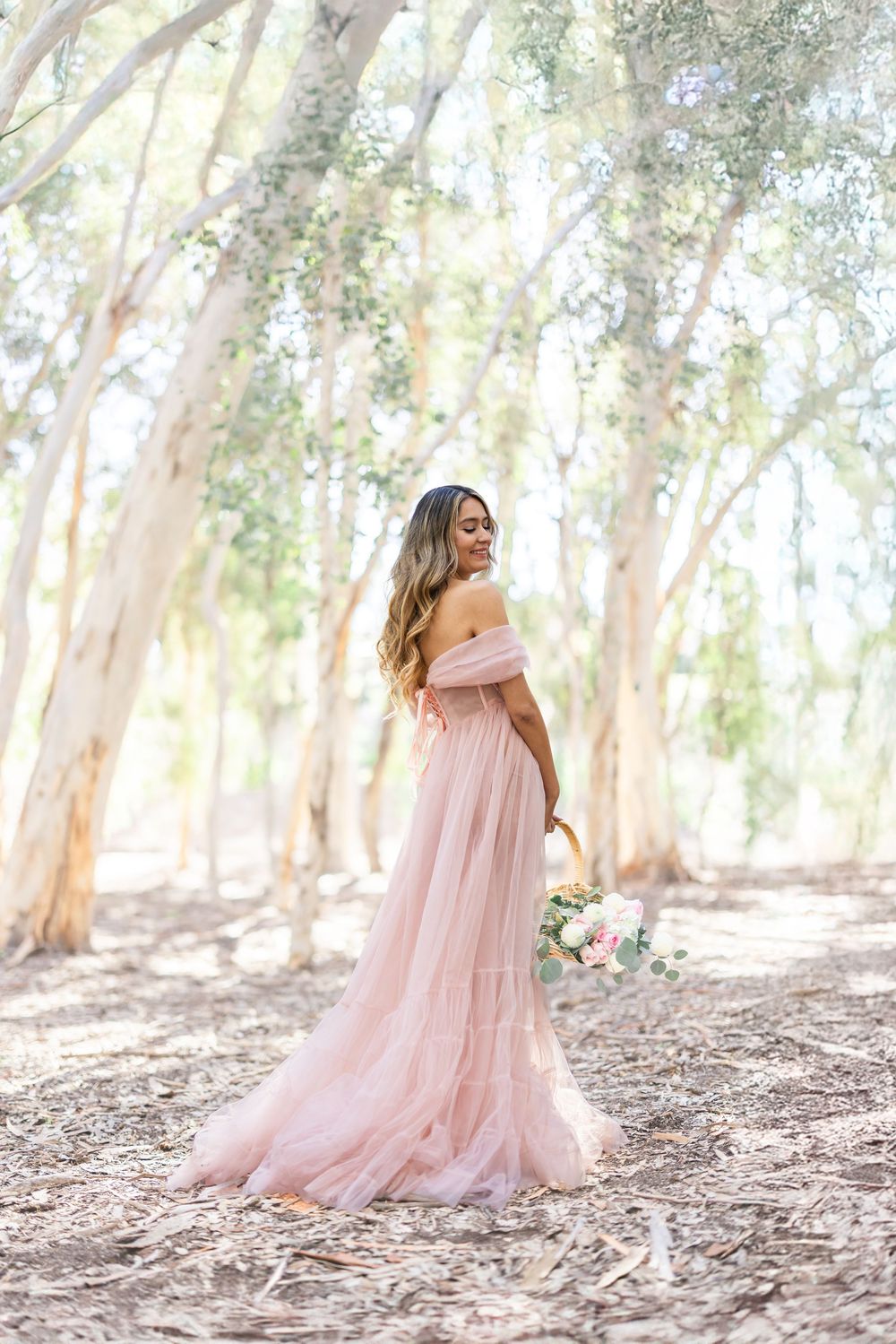Woman in blush pink off-shoulder tulle gown holding bouquet among sunlit eucalyptus trees.