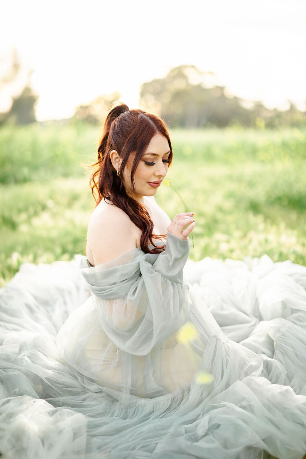 A woman with red hair in a flowing sage green tulle gown sits gracefully in a sunlit meadow.