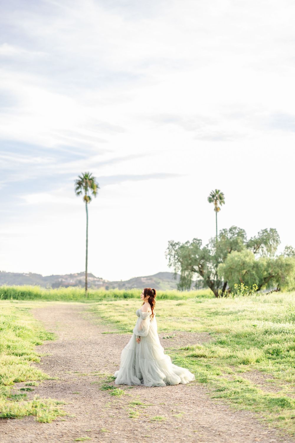 Bride in flowing white gown stands on dirt path surrounded by palm trees and green hills under soft cloudy sky.