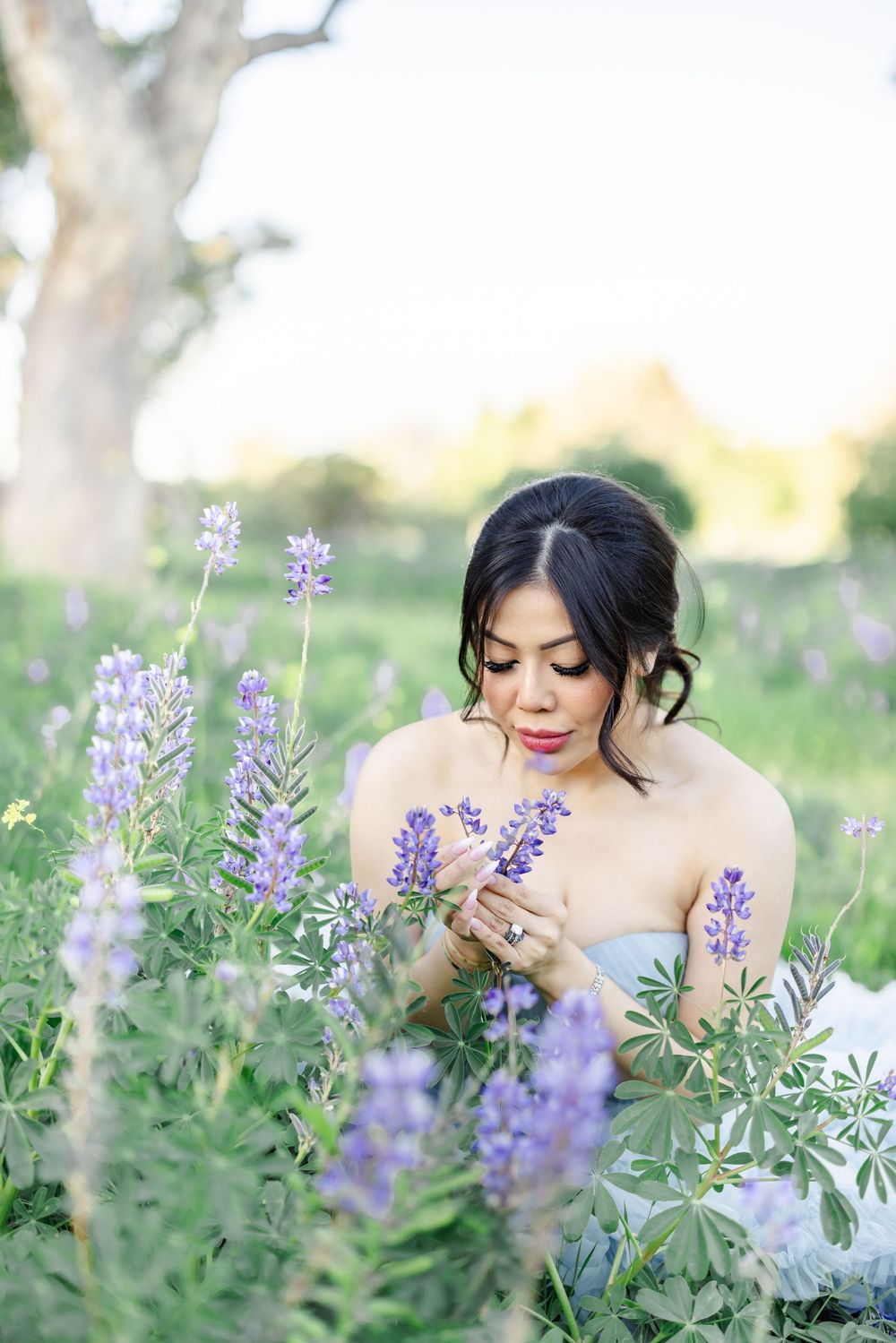 Woman in strapless gown sitting among purple lupine wildflowers in a sunlit meadow, gently holding blooms.