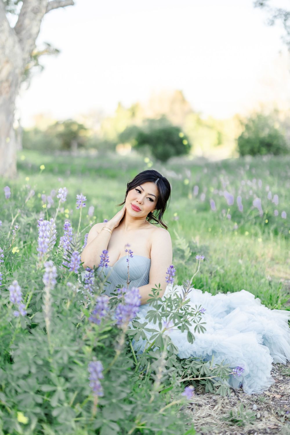Woman in gray gown sitting among purple lupine wildflowers in a lush green meadow during golden hour.